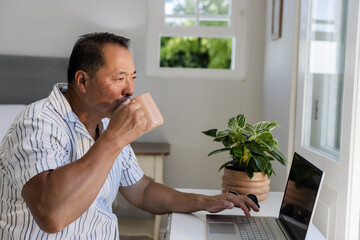 Senior Asian man drinking coffee while using laptop at home, enjoying morning routine