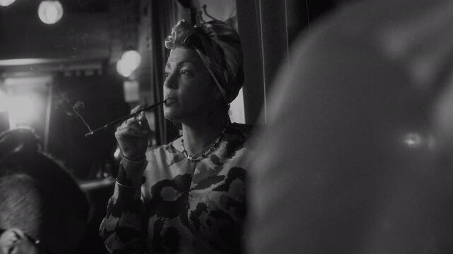 Bohemian Biracial woman in colorful attire elegantly smoking with cigarette holder in her hands while pianist playing nearby, giving cozy little live music performance at jazz lounge