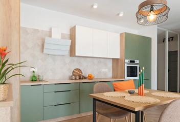 Fototapeta premium Modern kitchen interior with table and chairs. Green and white furniture with cabinet, oven and eaves and orange decoration. Design in new apartment. Real estate.