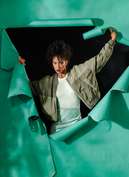 Woman breaking through green paper wall, wearing stylish jacket, looking determined