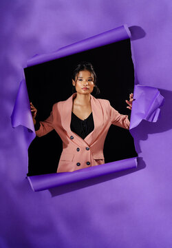 Confident Asian woman in stylish blazer emerging through torn purple paper