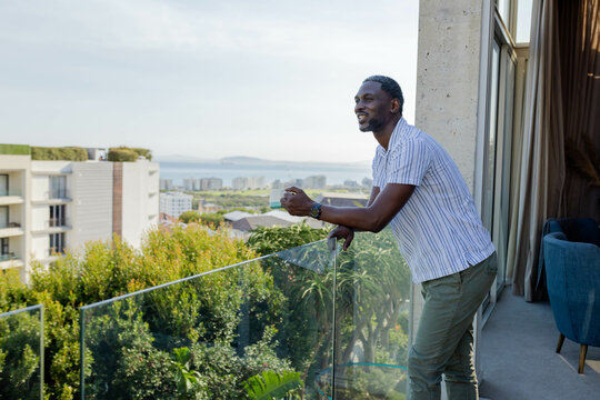 Smiling African American man enjoying morning view from balcony overlooking cityscape and greenery - Powered by Adobe