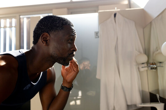 African American man grooming beard in bathroom, wearing tank top, focusing on reflection