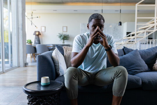 Thoughtful African American man sitting on sofa at home, contemplating with hands together