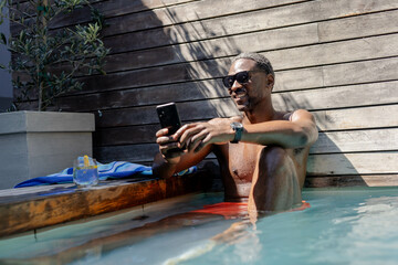 African American man relaxing in pool, using smartphone and enjoying sunny day