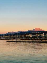 Calm sea reflecting warm hues of sunset sky, featuring Island harbor, breakwater, and majestic mountains rising in background, creating tranquil evening scene. Sunset light, rocky breakwater, Island