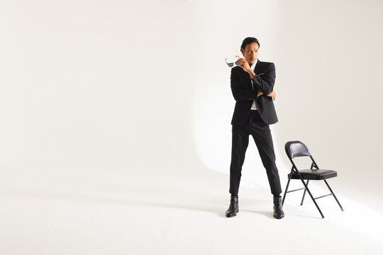 Confident man in stylish black suit posing with sunglasses near chair