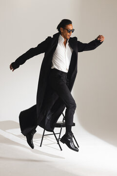 Stylish man in long coat and sunglasses confidently jumping off chair