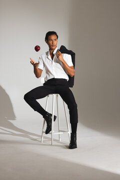 Stylish man in white shirt juggling apple while sitting on stool in studio