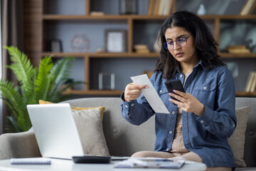 Upset young Indian woman sitting on sofa at home near table with laptop, holding phone in hands and...
