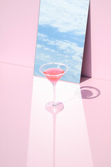  A glass with a drink placed in front of a mirror that reflects sky and clouds in the background. Summer aesthetic surreal composition. Beverage party concept.