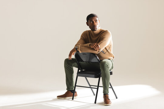 Stylish man in sweater sitting on chair, looking thoughtfully in studio - Powered by Adobe
