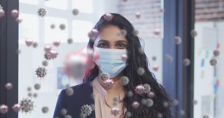 Image of covid 19 cells floating over portrait of biracial woman wearing face mask in office