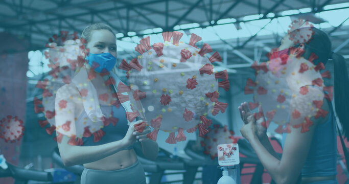 Covid-19 cells floating against two women wearing face masks sanitizing their hands at the gym