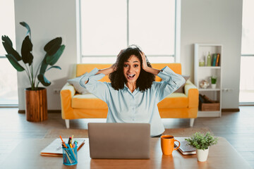 Young woman working on a laptop from home feeling cheerful and surprised, sitting in a stylish...