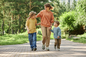 Obraz premium Full length front view of cheerful mother with two boys holding hands while walking towards camera in green park copy space