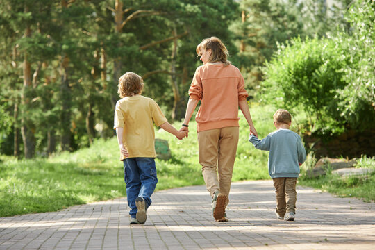 Full length back view of mother with two boys holding hands and walking away from camera in green park copy space