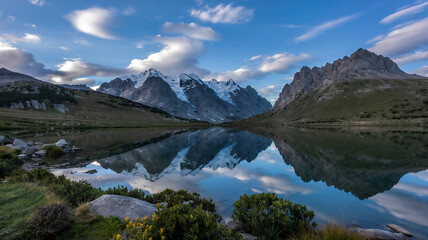 The clear, turquoise waters of the lake are surrounded by lush green slopes and rocky outcrops, with a few scattered wildflowers adding bursts of color.
