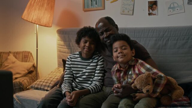 Portrait of loving African American dad snuggling on couch with his sons, smiling and looking at camera together