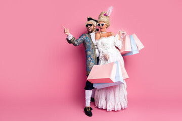 Joyful Couple in Elegant Period Costumes Carrying Shopping Bags Against a Pink Background