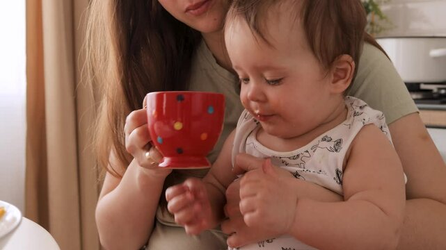 A young child sits in a high chair, curiously sipping from a bright red cup while being held by their mother. The cozy setting is filled with warm lighting and a relaxed atmosphere
