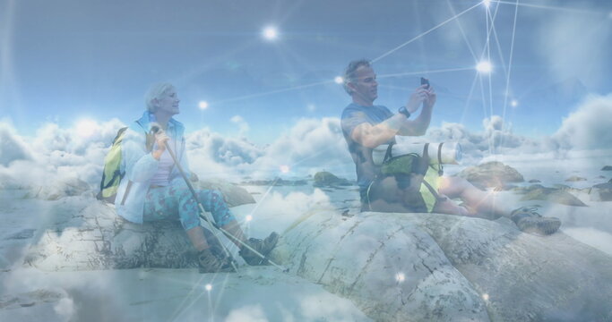 Caucasian senior couple hiking, taking photo with smartphone, over clouds and network of connections