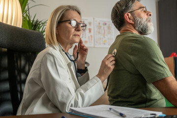 Female doctor using stethoscope to listen to male patient's lungs during medical examination in...