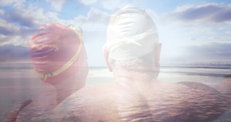Image of glowing light over happy senior couple in swimming hats by seaside