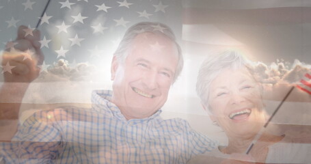 Image of glowing light over portrait of happy senior couple waving american flags