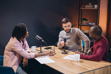 Three podcasters engaging in conversation while recording their latest episode at a wooden table in...