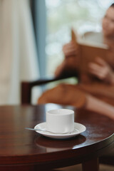 A serene indoor scene featuring a white cup on a wooden table, with a blurred background of a person reading a book, depicting a peaceful coffee break