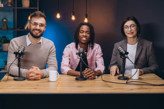 Three podcasters sitting at a table with microphones and mugs, recording a new episode in a professional studio