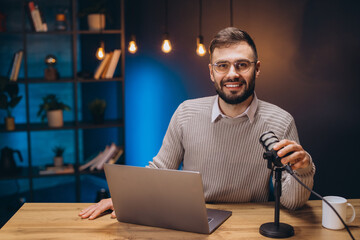 Happy male podcaster speaking into a microphone while recording audio podcast in home studio with...
