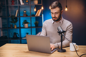 Young man recording a podcast in a professional studio, using a laptop and a microphone, with a bookshelf in the background © anatoliycherkas