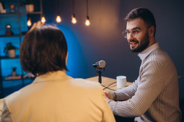 Two podcasters sitting at a table and talking into a microphone, recording a new episode for their...