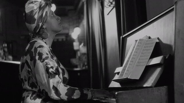 Side view medium black and white shot of cinematic woman in patterned outfit and headwrap playing piano passionately, surrounded by quiet elegance of vintage bar