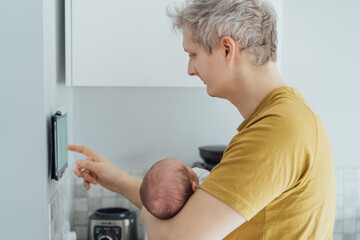 Young man with newborn baby adjusts settings, switch on wall-mounted thermostat or smart control panel, managing temperature, air conditioning system. Modern technology, controlled home environment