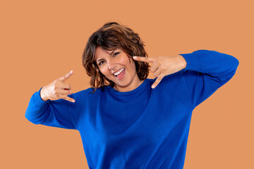 Joyful woman expressing excitement in blue sweater against orange background while making rock and...
