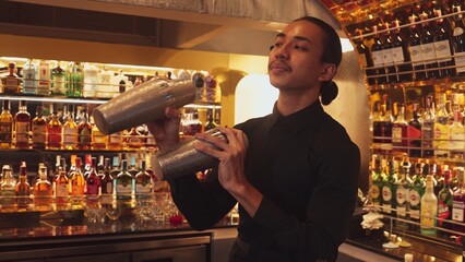 Smiling Asian smart bartender shaking with two hands in mixed cocktail in shaker preparing at luxurious counter bar, preparation special refreshment alcohol drink nightlife warm nightclub. Vinosity.
