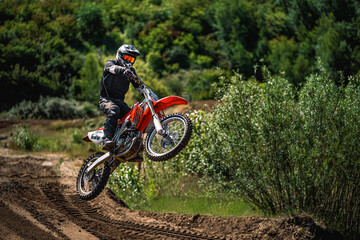 Motocross rider jumping off-road on a dirt track surrounded by lush greenery. The athlete is fully equipped with protective gear, helmet, and goggles. The image captures the thrill, speed, and energy 