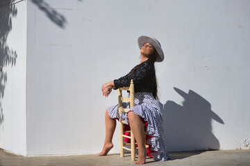 Young woman, beautiful, brunette, flamenco dancer, wearing a beautiful dress with jacket and hat, barefoot, posing sitting on a chair casting her shadow on a white wall.