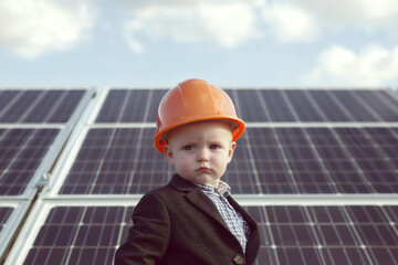 A baby wearing a suite and hard hat standing in front of a big solar panel