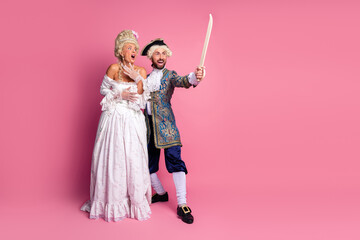 Elegant couple in historical attire posing on pink background depicting a lively and humorous...