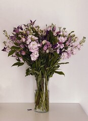 A bouquet of pink and purple flowers with lush green leaves is arranged in a clear glass vase. The vase is set against a plain white background, highlighting the vibrant colors of the flowers. 