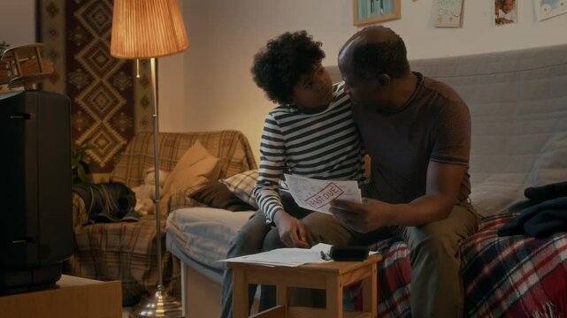 African American man, going through tough times, hugging his son, while looking at past due bills in cozy living room