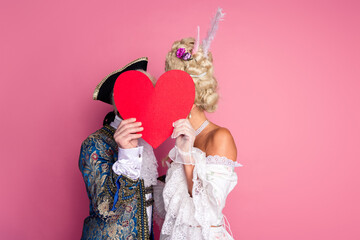 Elegant couple in historical costumes holding a heart on a pink background, perfect for romantic or...