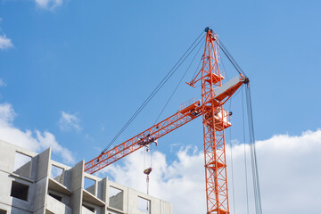 Tall orange construction crane towers over a building site, against a backdrop of a bright blue sky with clouds.
