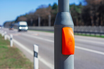 Orange pedestrian crossing button on a pole stands along a highway with a truck approaching in the...