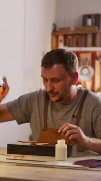 Leathersmith adjusts table lamp for comfortable leather processing in workshop. Bearded master crafts item in small studio. Workplace of handcraft designer