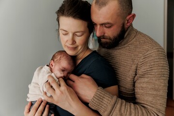 A couple lovingly holds their newborn baby in a cozy indoor setting. The mother gazes at the baby...